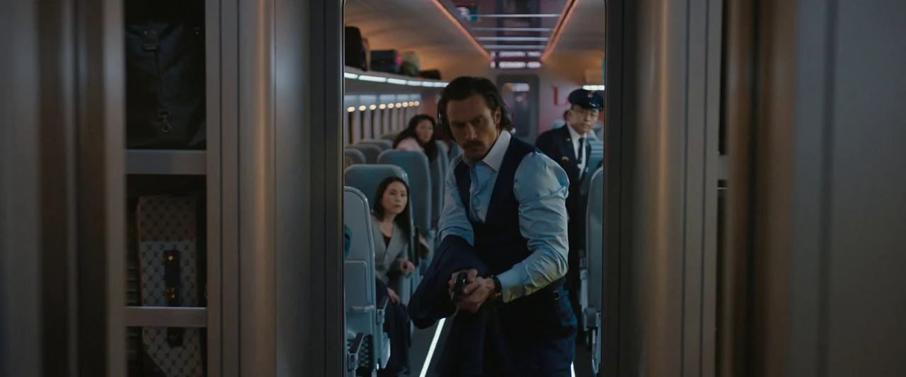 Bullet Train (2022) movie still: medium — A modern train car cabin features rows of blue seats and luggage racks above. A man walks through the…