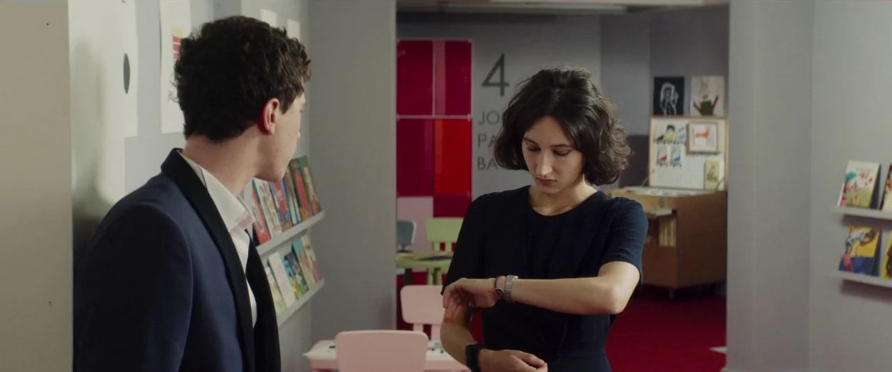 Nocturama (2016) movie still: medium — A modern building interior with pale gray walls, shelving units, and children's furniture. A young woman…