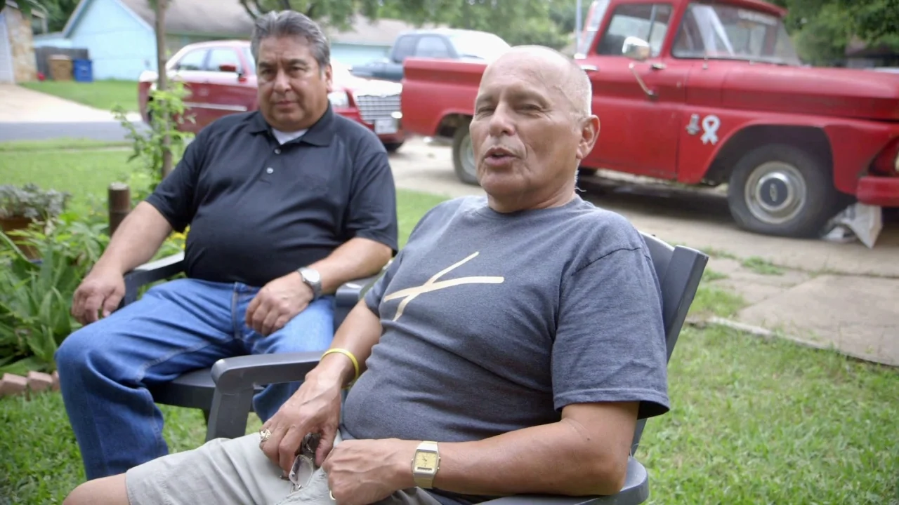 Tower (2016) movie still: medium — Two middle-aged men sit in plastic chairs on a residential lawn. A bright red vintage pickup truck is parked in…