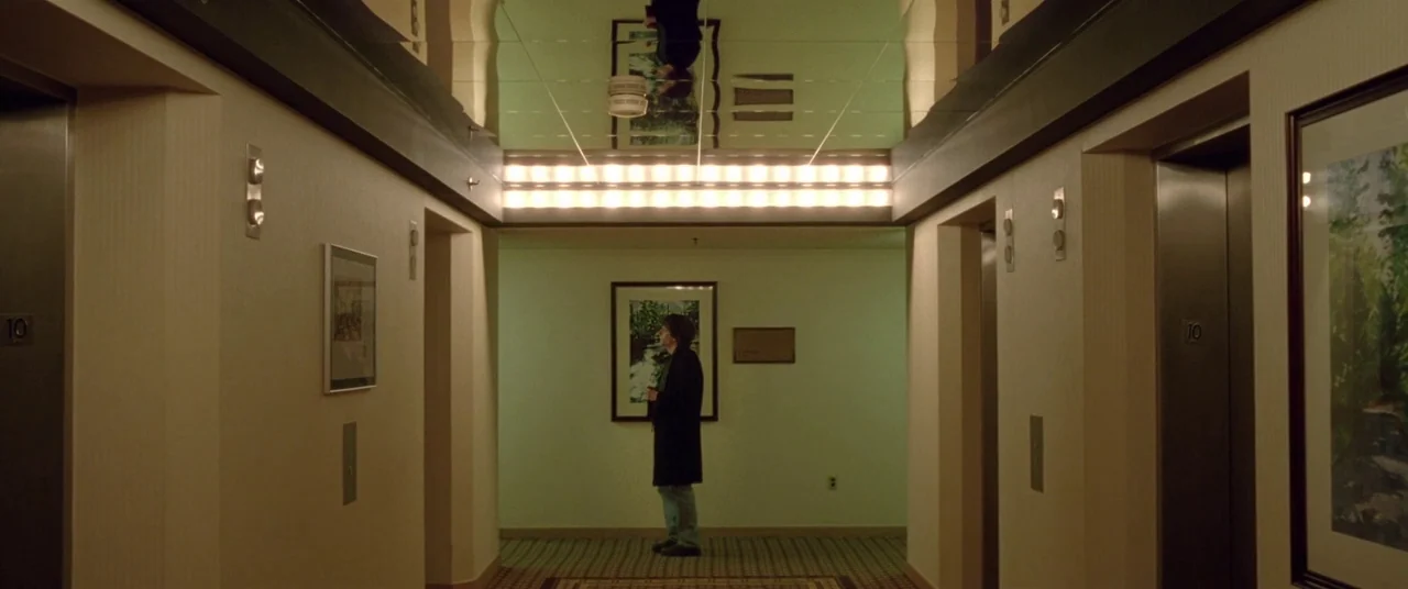 The End of the Tour (2015) movie still: wide — A symmetric hotel elevator lobby features beige walls, reflective mirrored ceiling panels, and framed…