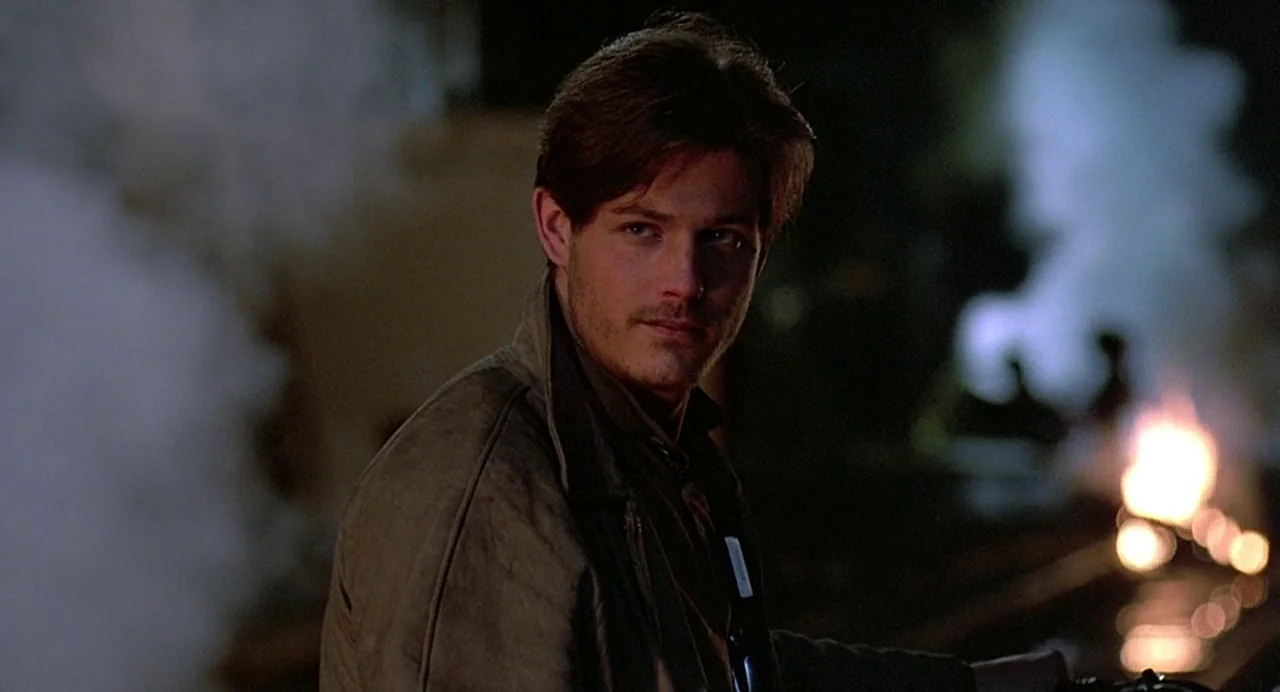 Streets of Fire (1984) movie still: medium — A man stands outdoors near railroad tracks at night in a steam-filled environment. He gazes off-camera…