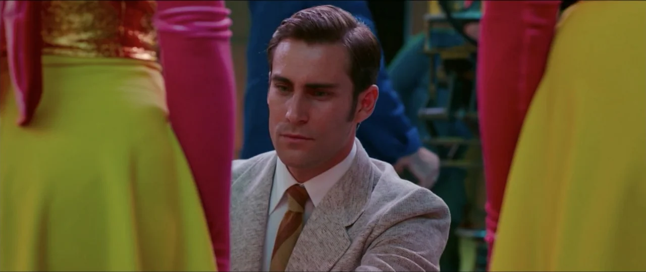 My Heart Goes Boom! (2020) movie still: medium — A man in a suit stands between two individuals wearing vibrant yellow skirts. The setting features…