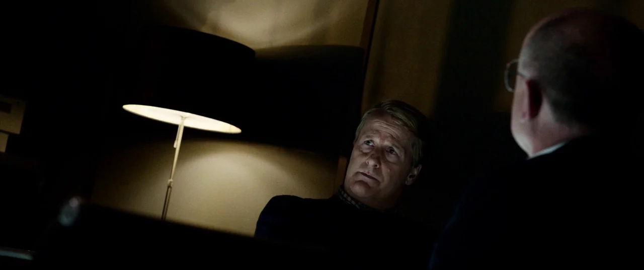 Steve Jobs (2015) movie still: medium — A dark hotel room with a single lamp casting light on a seated man. An older man in the foreground is seen…