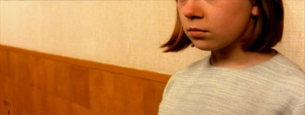 Carne (1991) movie still: closeup — A plain interior corridor features a textured white wall and a brown wood-paneled lower section. A young girl…