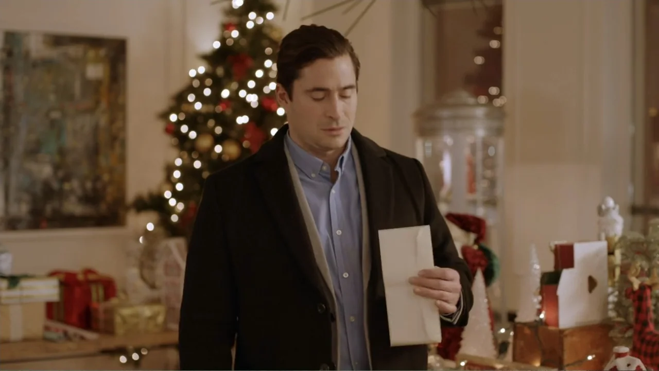 Loving Christmas (2022) movie still: medium — A man stands in a festive holiday decorated space with a Christmas tree behind. He holds a white…