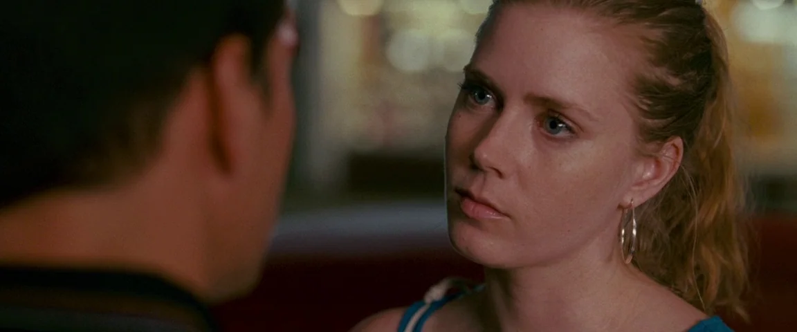 The Fighter (2010) movie still: closeup — A woman looks intently at an interlocutor inside a dimly lit indoor establishment. The background is…