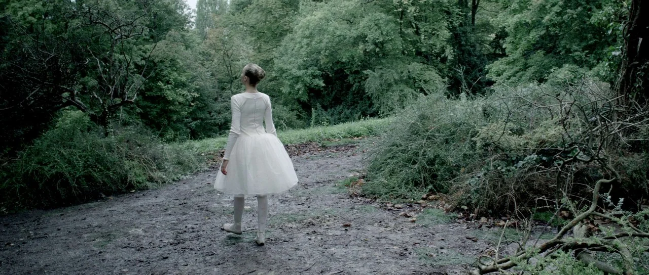 Livid (2011) movie still: fullbody — A dense, overgrown forest area features a narrow, mud-caked dirt path. A woman in a white ballerina dress…