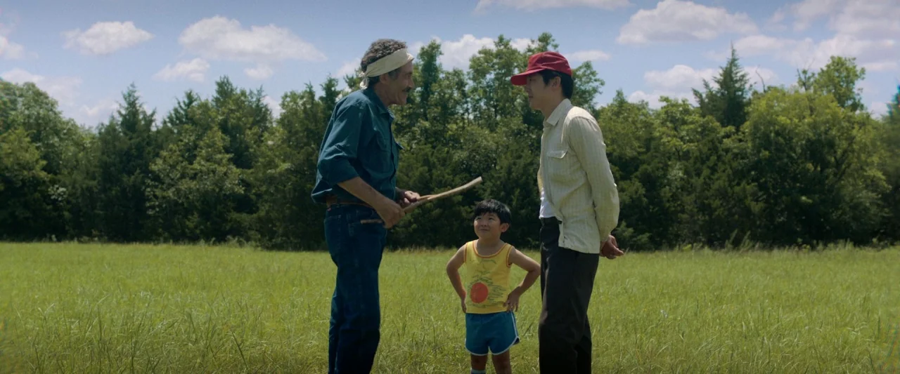 Minari (2020) movie still: fullbody — A sunlit green meadow stretches toward a dense tree line under a cloudy sky. An older man, a young boy, and a…