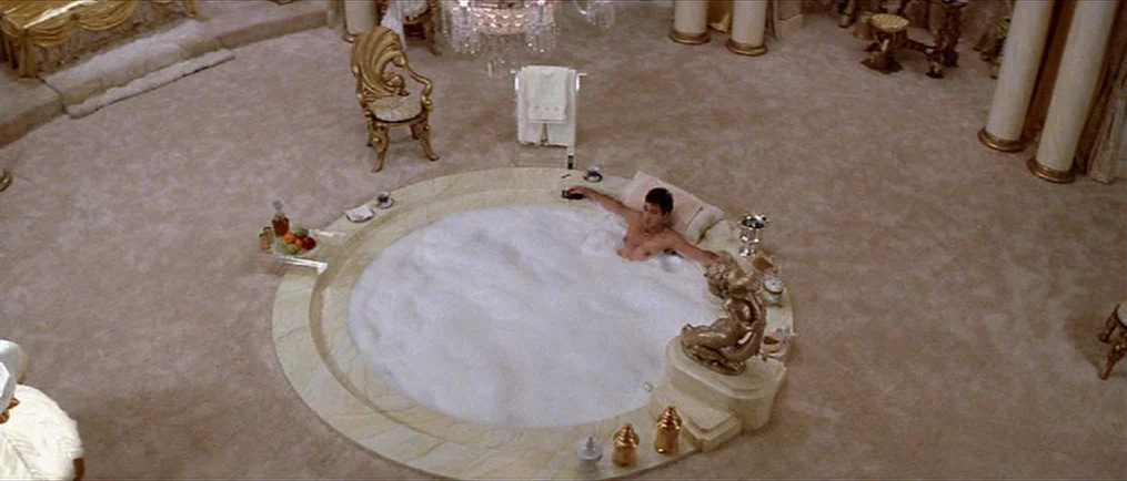 Scarface (1983) movie still: wide — A lavish 1980s circular bathroom with wall-to-wall carpeting and ornate gold accents. A man relaxes in a large…