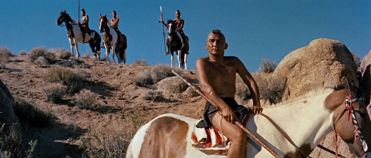 Comanche Station (1960) movie still: wide — A dry, rocky hillside under a clear blue sky serves as a backdrop. Four men with mohawks ride spotted…