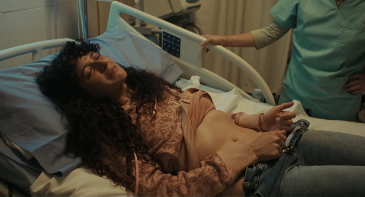 Belén (2025) movie still: medium — A clinical space contains a white adjustable medical bed with blue bedding. A woman in a patterned hoodie rests…