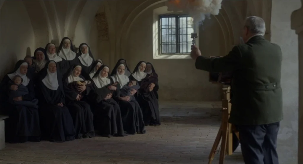 The Innocents (2016) movie still: wide — A stone-walled interior with arched ceilings and a dirt floor holds a group of nuns. The photographer…
