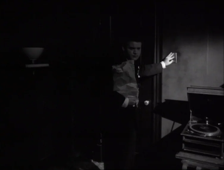Eraserhead (1977) movie still: medium — A dark room features a door and a record player on a small wooden stand. A man in a suit enters the space…