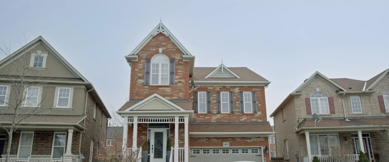 Motherly (2022) movie still: establishing — A row of three detached suburban brick houses stands under an overcast sky. The central house features a…