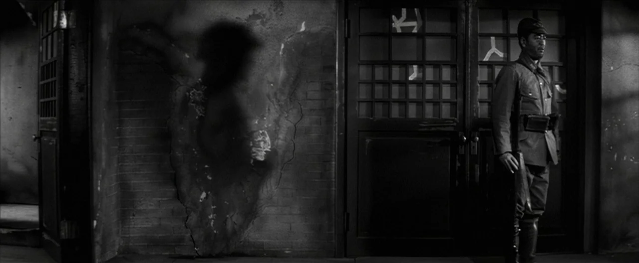 Story of a Prostitute (1965) movie still: medium — A dark urban building exterior features a damaged brick wall with a scorched marking. A soldier…