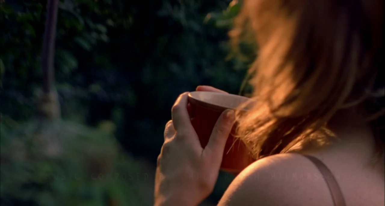 In The Cut (2003) movie still: closeup — A person holds a red mug in a dimly lit, foliage-filled outdoor environment. Warm sunlight catches the hair…