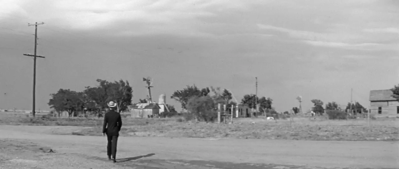 Hud (1963) movie still: wide — A flat, sparsely populated rural landscape contains several wooden structures and a metal windmill. A lone man in a…