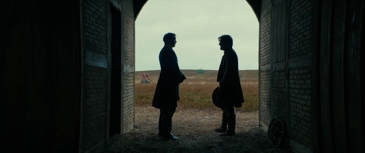 The Promised Land (2023) movie still: fullbody — A dark brick barn entrance frames a wide view of a desolate, grassy field. Two men stand in…