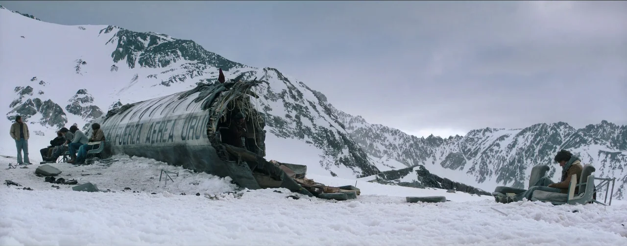 Society of the Snow (2023) movie still: wide — The torn fuselage of a small airplane rests on a steep, snow-covered mountain slope. Several people…