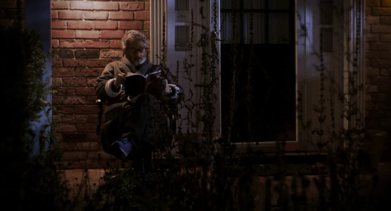 Happer’s Comet (2022) movie still: medium — An elderly man reads a book while sitting on a porch at night. A red brick wall and white front door…