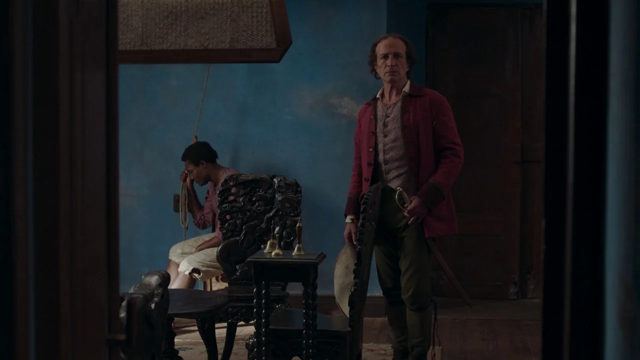 Zama (2017) movie still: medium — An 18th-century parlor with distressed blue walls features dark ornate wooden furniture. A man stands in the…