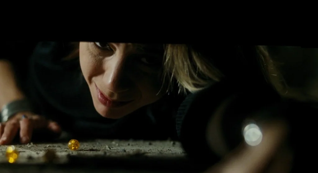 Mimic (1997) movie still: closeup — A person lies on a dusty ground surface in a dark environment. The woman smiles while looking forward,…