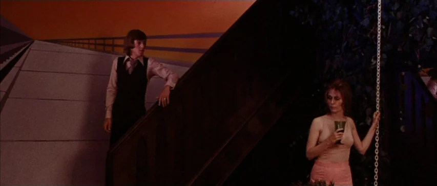 Messiah of Evil (1973) movie still: wide — A living room interior features a large wooden staircase and a mural on the wall. A young man stands by…