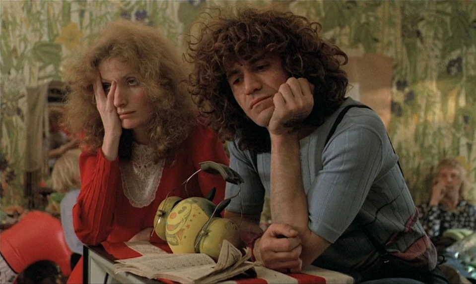 Lions Love (1969) movie still: medium — A room features patterned floral wallpaper and a table where an old clock sits. A man and a woman sit…