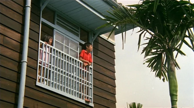 A Summer at Grandpa’s (1984) movie still: medium — A wooden building features an elevated balcony with a white latticed railing. Two children lean…