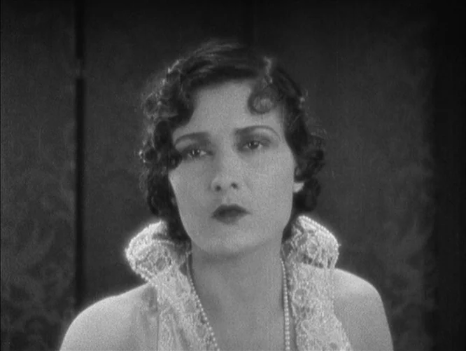 The Last Command (1928) movie still: closeup — A woman stands in a softly lit interior space with a dark patterned backdrop. She gazes directly at…