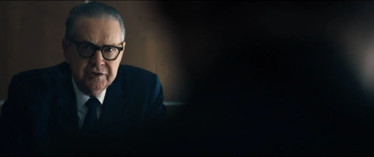 First Man (2018) movie still: medium — A dimly lit professional office with a dark wooden wall panel background. An older man in a suit sits and…