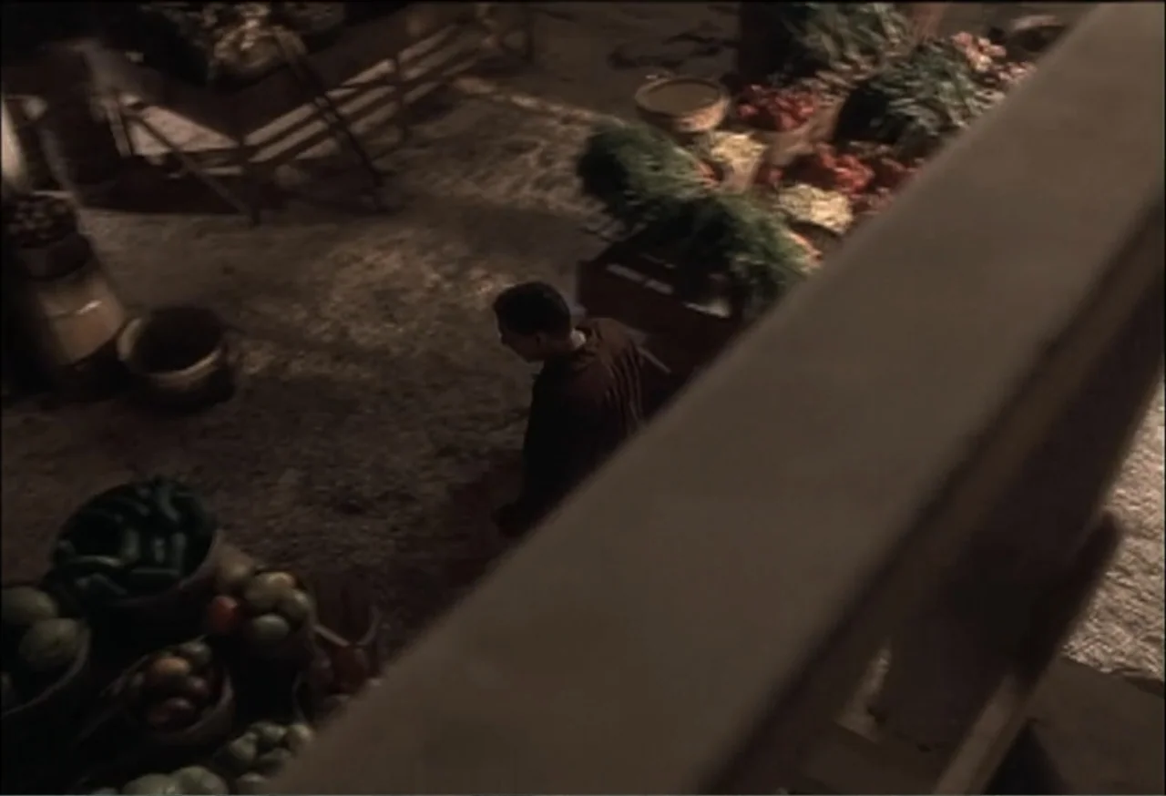 Vendetta (2022) movie still: wide — An outdoor night market with wooden crates filled with produce and straw flooring. A lone male figure walks…
