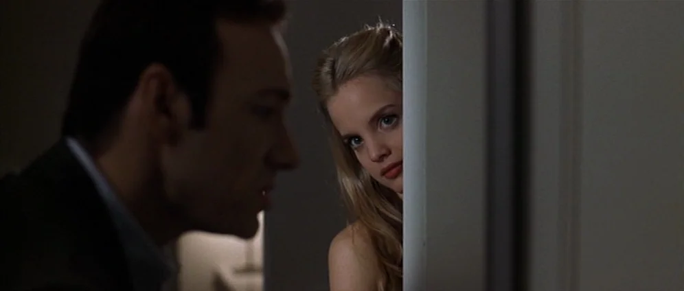 American Beauty (1999) movie still: medium — A dark, minimally furnished space is divided by a white vertical wall partition. A young woman peeks…