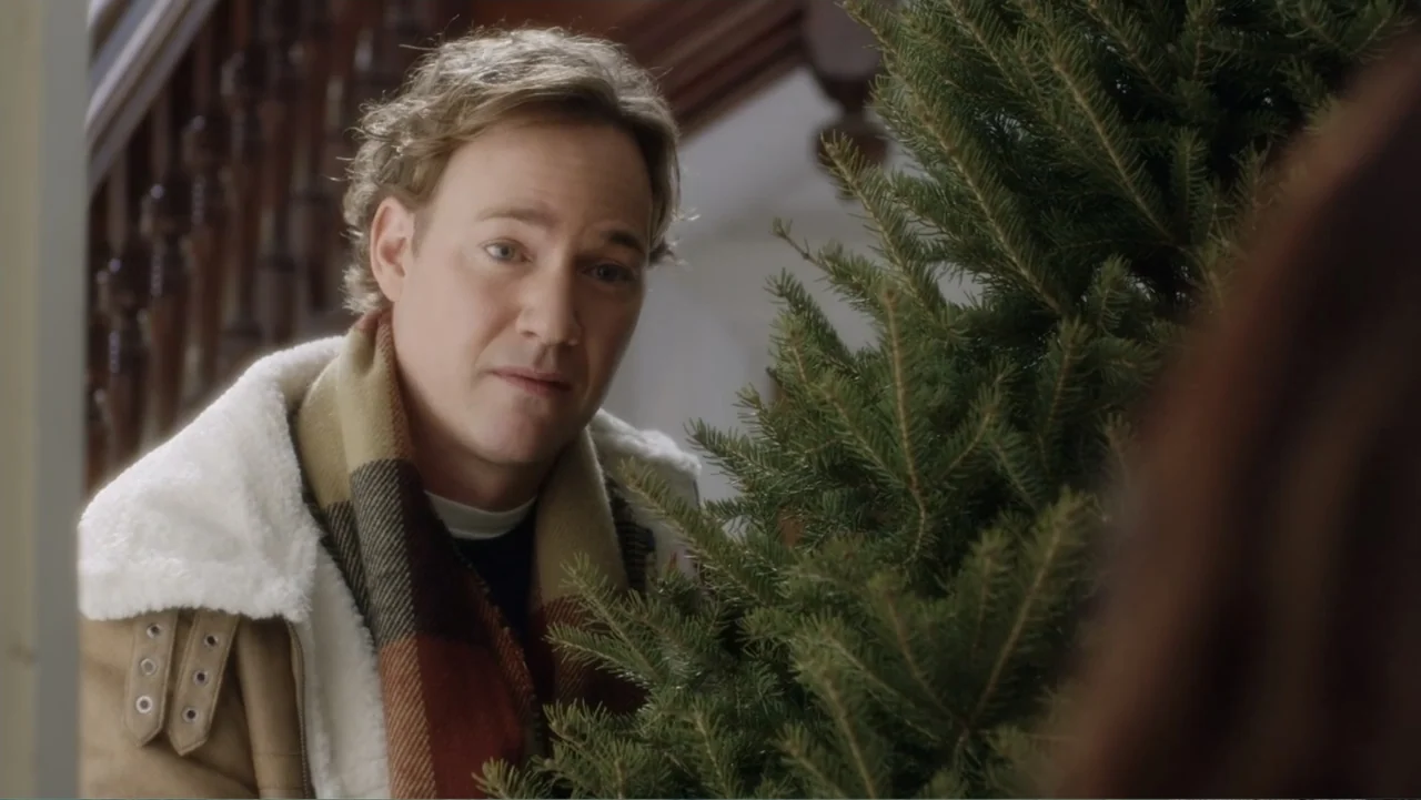 A Christmas Break (2020) movie still: medium — A residential foyer features a staircase and a large potted evergreen tree. A man in a shearling coat…
