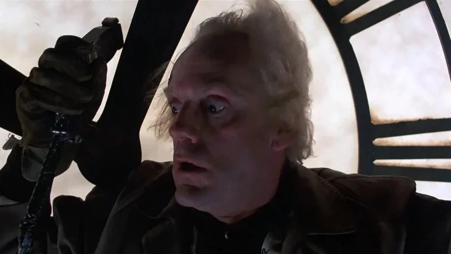 Back to the Future (1985) movie still: closeup — An elderly man in a dark coat grips a pole inside a large clock structure. The illuminated glass…
