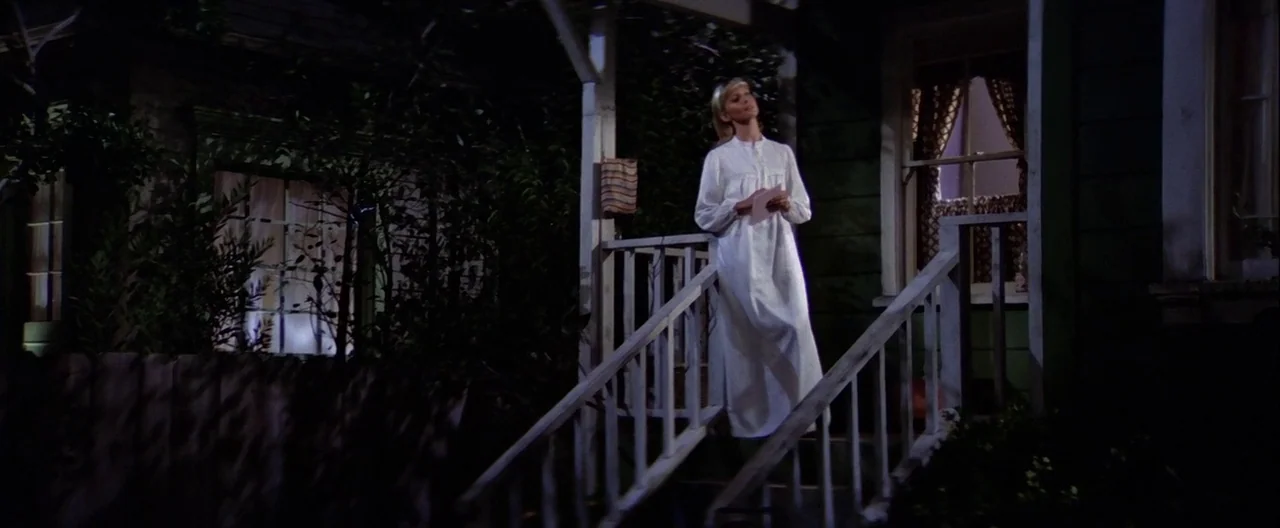 Grease (1978) movie still: fullbody — A wooden porch at night illuminated by light spilling from inside a house window. A woman in a white nightgown…