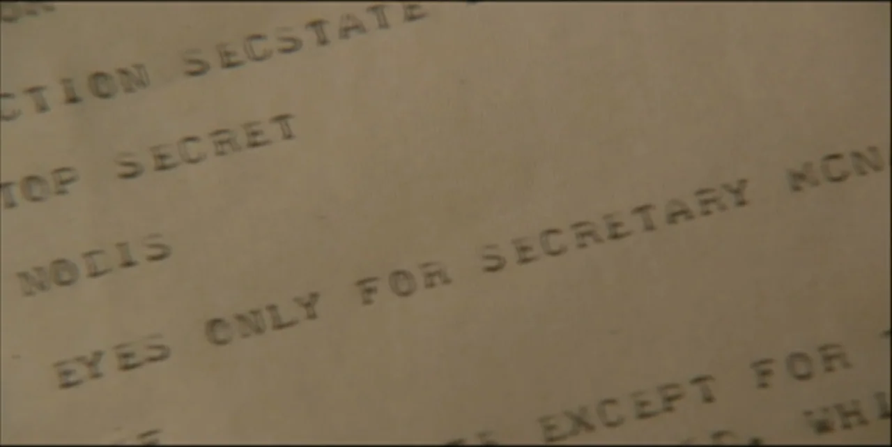 Path to War (2003) movie still: closeup — A close-up view of an aged, off-white document featuring stamped typewriter text. The document bears…