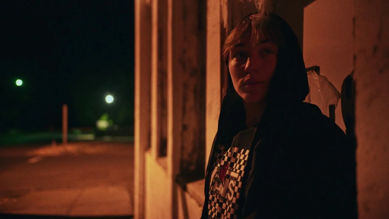 Gasoline Rainbow (2023) movie still: medium — A person stands at the edge of a building exterior during the night. The surrounding dark street shows…
