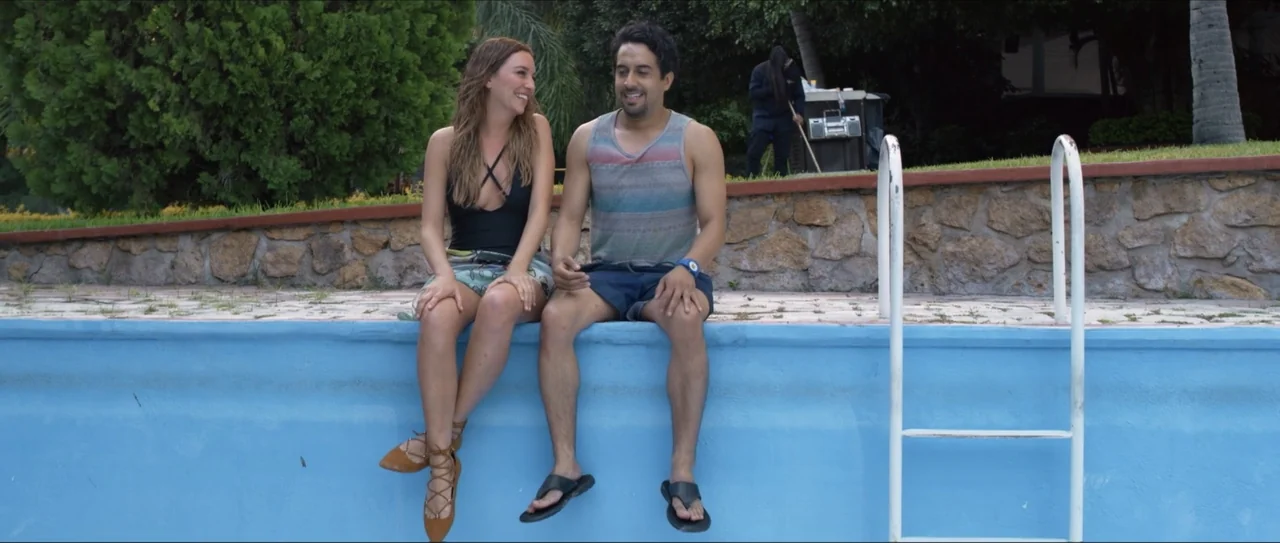 Mirreyes vs Godínez (2019) movie still: medium — An outdoor concrete pool edge sits before a stone wall and green trees. A man and a woman sit on…