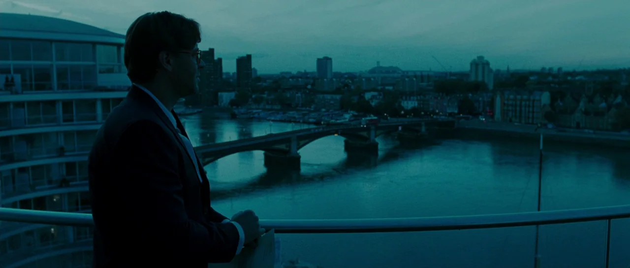 A Good Year (2006) movie still: medium — A man stands on a high-rise balcony at twilight overlooking a wide river. The city skyline and a bridge are…