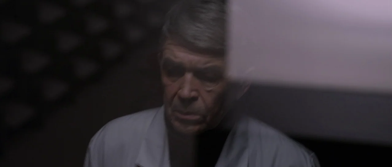 Vanishing Waves (2012) movie still: closeup — An older man in a white coat is shown in a dimly lit space. He looks downward with a neutral…