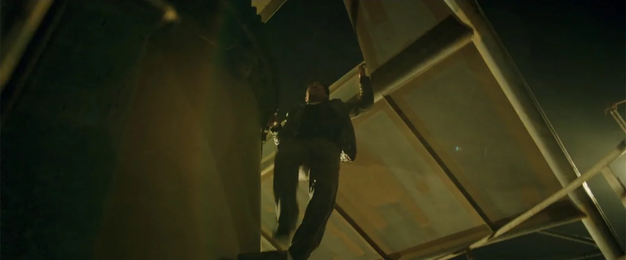 Skiptrace (2016) movie still: medium — A dark night scene at a construction site with exposed steel beams overhead. A man hangs from a metal beam…