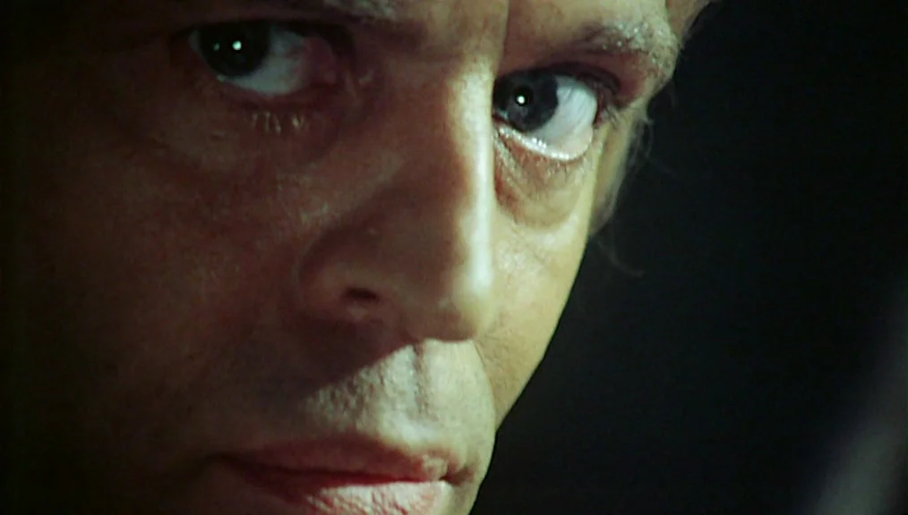 The Great Silence (1968) movie still: closeup — A dimly lit interior space with a pitch-black background. A man stares directly toward the side of…
