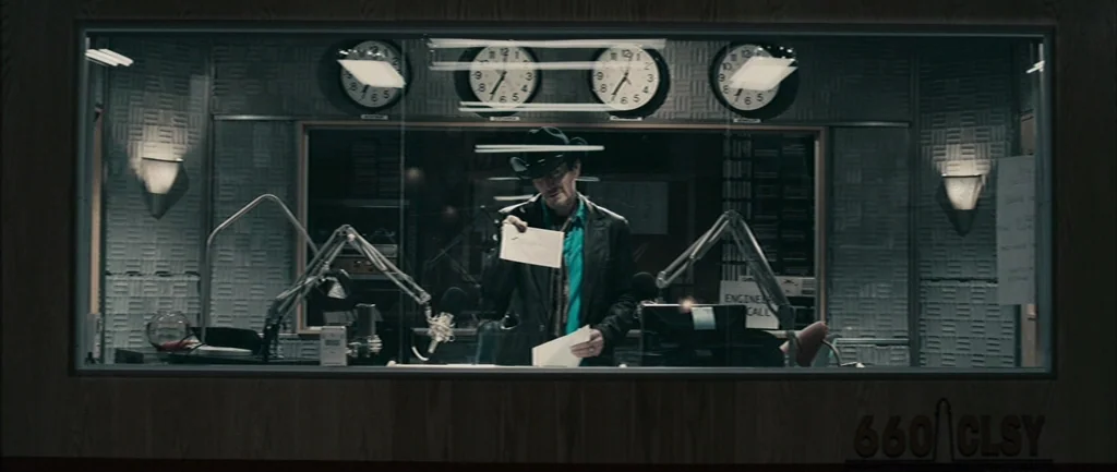 Pontypool (2008) movie still: medium — A sterile radio broadcast booth features sound-dampening wall panels and a row of clocks. A man in a hat…