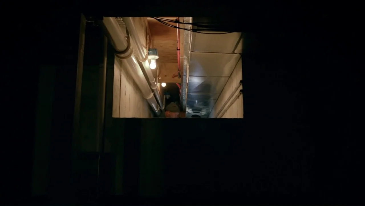 Clear History (2013) movie still: wide — An unfinished crawlspace area features exposed wooden beams, pipes, and metal ductwork. A single…