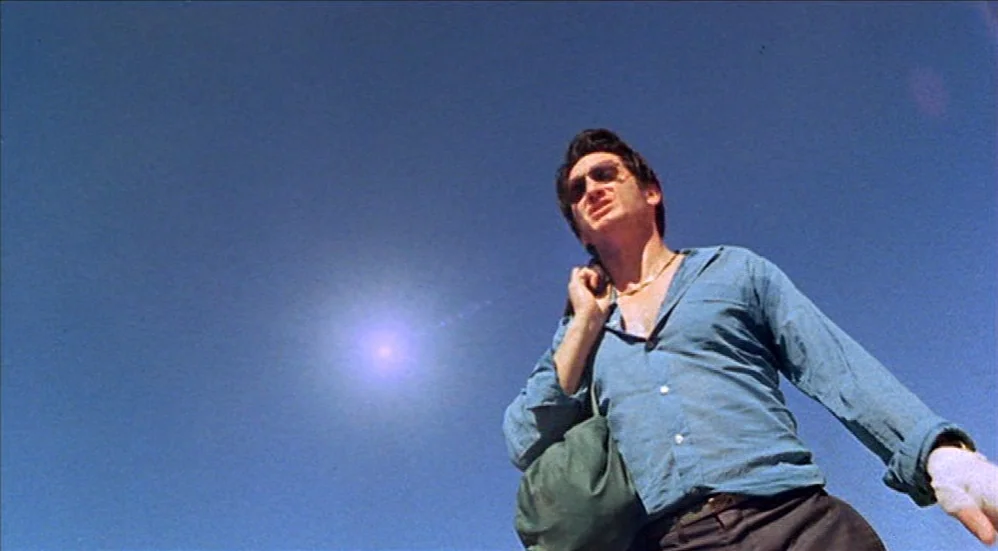 U-Turn (1997) movie still: medium — A man walks against a clear, vibrant blue sky in bright daylight. He carries a dark bag over his shoulder while…