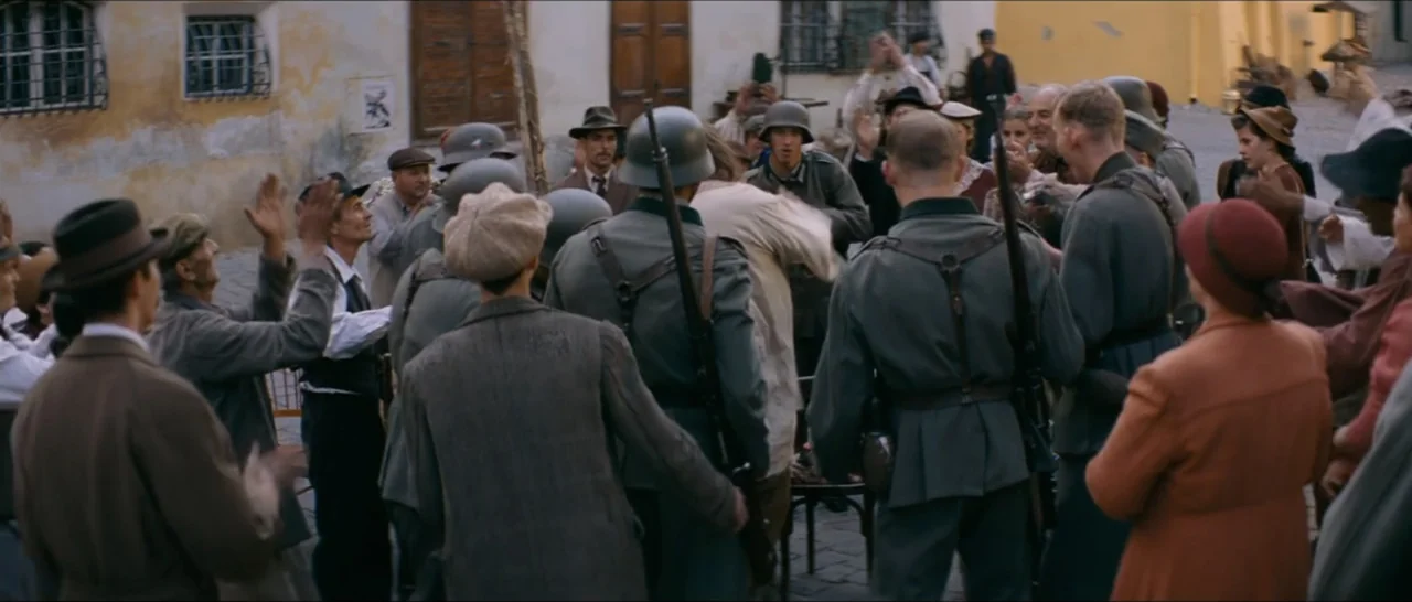 A Farewell to Fools (2013) movie still: wide — A crowd of civilians in 1940s attire gathers in a European cobblestone village square. Soldiers in…