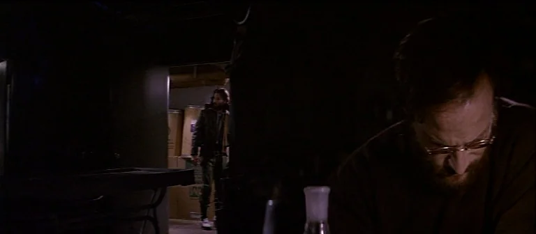 The Thing (1982) movie still: medium — A dimly lit basement features stacked cardboard boxes and scientific glassware on a table. A bearded man…