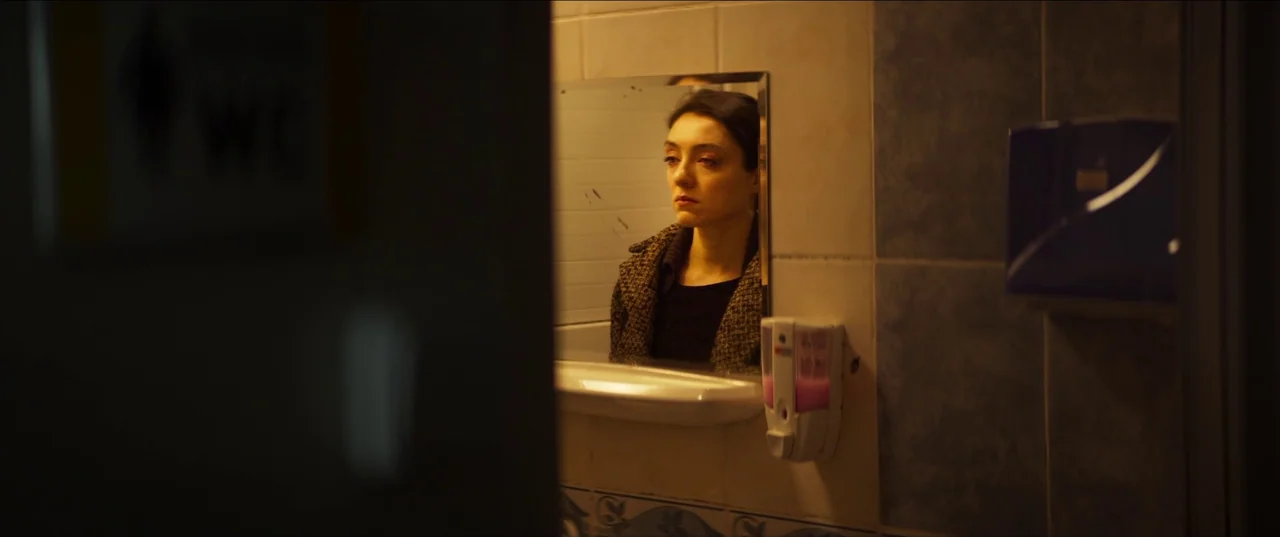 About Dry Grasses (2023) movie still: medium — A tiled public restroom with a mounted soap dispenser next to a wall mirror. A woman stares at her…
