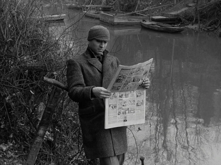 Band of Outsiders (Bande à part) (1964) movie still: medium — A man stands on a muddy bank beside a calm, reflective river. He leans against a…